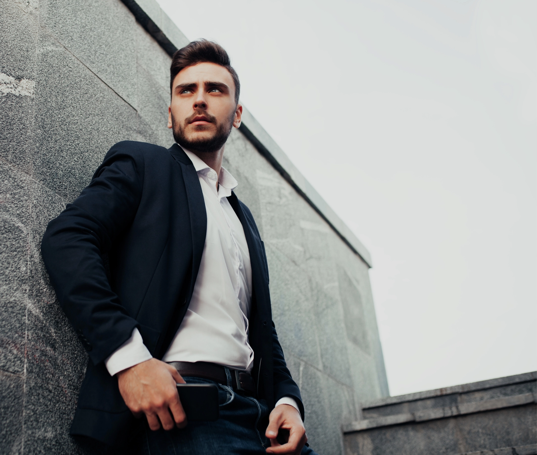 Man in a suit leaning against a concrete wall, hand in pocket, holding a smartphone. - Bio, Webster, TX