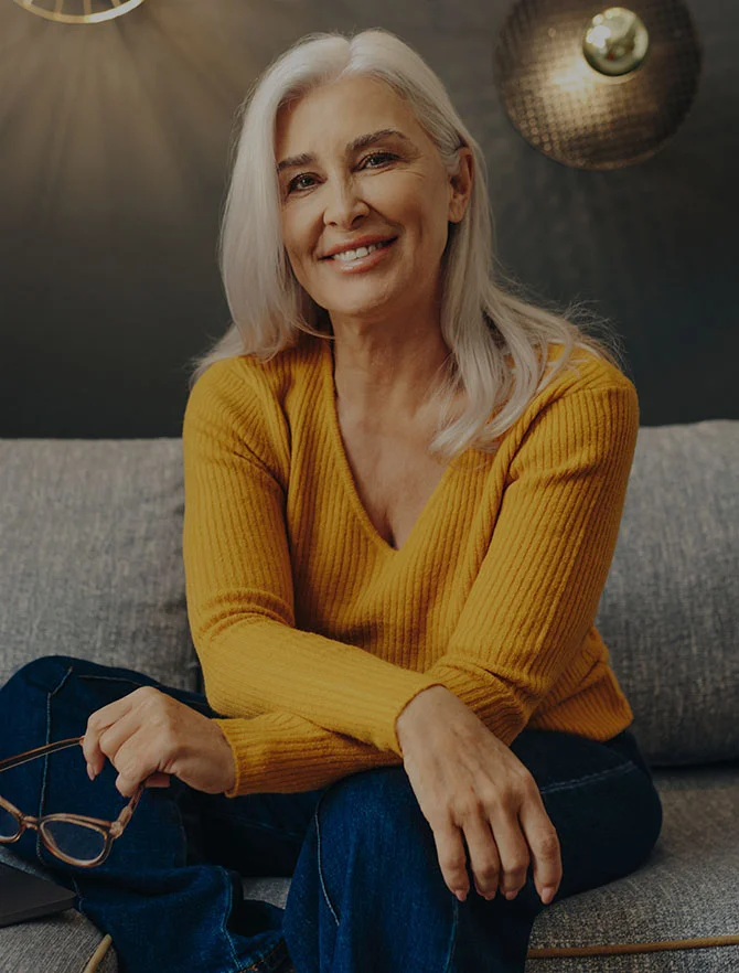 Middle aged women with grey long hair in a yellow sweater and blue jeans sitting on a gray couch holding eyeglasses. - Procedures in Webster, TX