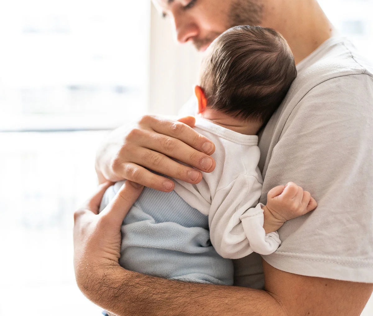 A close-up shot captures a baby nestled comfortably in his father's arms, with the father's hand gently patting the baby's back. The baby, wearing a white onesie and soft blue pants, is the focal point of the image, conveying a sense of warmth and security. The father, partially out of frame, wears an off-white T-shirt, adding to the cozy, tender atmosphere of the scene. - Hydrocelectomy Treatment in Webster, TX