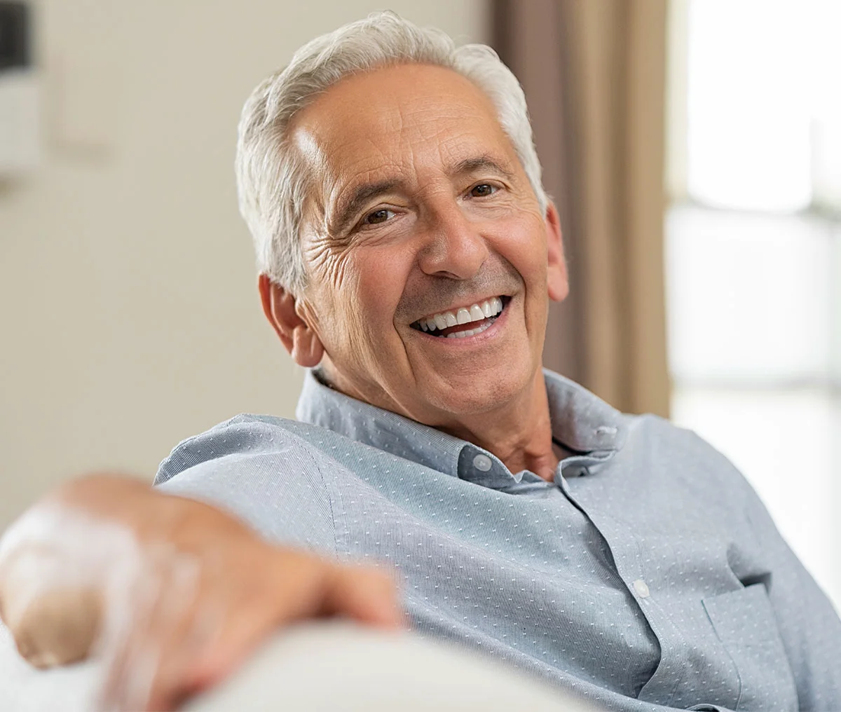 Semi close-up of a middle-aged man with white hair, wearing a light navy button-down long-sleeve shirt. He has his arm on the arm of the couch and is looking into the camera smiling. - Urolift System Alternatives in Webster, TX