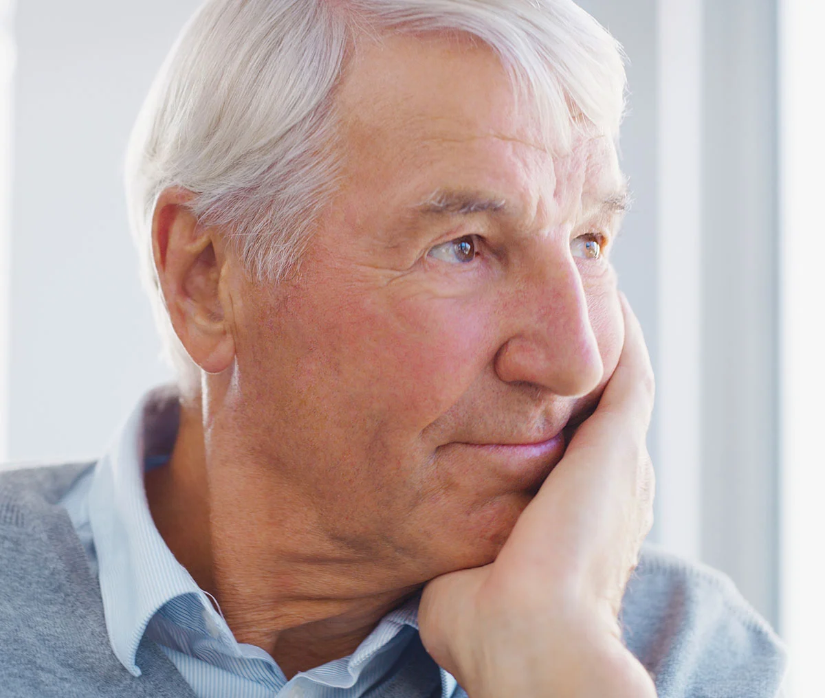 A close-up shot of a middle-aged man with brown eyes and white hair gazing thoughtfully out of a window, his face gently resting on his left hand. He has a subtle smile, giving him a warm and introspective expression. He’s dressed in a grey turtleneck sweater layered over a white and blue striped collared shirt, adding a touch of classic style to his look. - Aquablation in Webster, TX