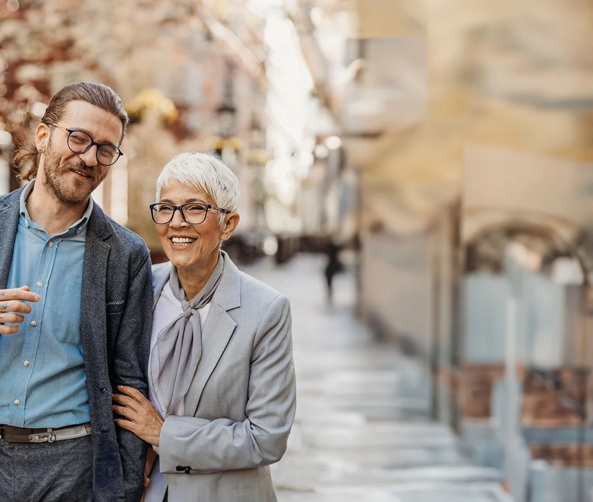 A younger man and an older woman walk side by side in a downtown setting, with the background gently blurred to emphasize their connection. The younger man on the left has brown hair pulled back into a ponytail, a beard, and wears black-framed glasses. He’s dressed in a blue button-down shirt under a dark grey blazer, with tattoos visible on his forearm and hand as he gestures while speaking. The older woman on the right, with short white hair and black-framed glasses, smiles warmly as she holds his arm closely. She’s dressed in a light grey blazer over a white shirt, complemented by a light grey scarf, exuding a sense of warmth and camaraderie between them. - Carrier Screening for Family Treatment in Webster, TX