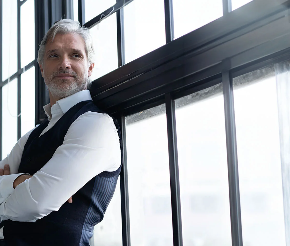 A middle-aged man with white hair and a beard leans against a wall full of windows. He is wearing a white long-sleeve button-down shirt and a navy suit vest. With his arms crossed, he looks past the camera, conveying a thoughtful or confident expression. - Kidney Stone